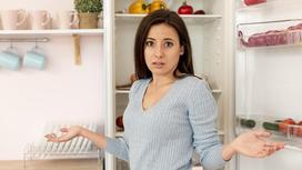 Девушка стоит перед открытым холодильником с продуктами и разводит руками