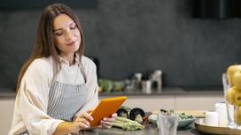 Девушка стоит на кухне возле стола с продуктами и смотрит в планшет