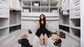 Девушка сидит в гардеробной и разводит руками. На полу расставлены разные пары обуви