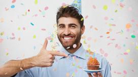 Бородатый молодой мужчина в праздничном колпачке улыбается и показывает пальцем на маленькое пирожное с одной свечой
