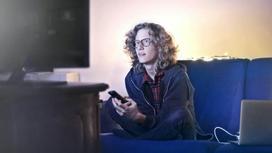 Молодй человек в очках сидит на диване и смотрит телевизор