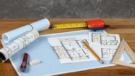 На столе развернута миллиметровая бумага с техническим планом дома. Рядом лежит рулетка, треугольник, карандаш, канцелярский нож, бумажные свертки
