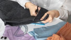 Темные джинсы разреазают ножницами на части. На столе лежат цветные лоскуты ткани, транспортир, карандаш