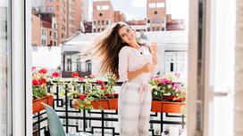Девушка стоит на отурытом балконе и улыбается. В балконных ящиках цветет красными и розовыми цветками пеларгония
