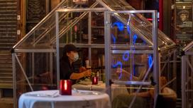 женщина в маске сидит в стеклянном домике в ресторане