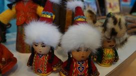 Две куклы в женских кахаских национальных костюмах и одна фигурка в мужском казахском национальном костюме. Фигруки стоят на столе рядом с другими экспонатами выставки