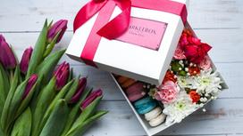 Открытая подарочная коробка с цветами и пирожными внутри. Рядом лежит букет тюльпанов