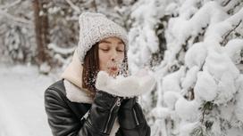 Девушка в вязаной шапке, дубленке и варежках держит в руках снег и дует на него