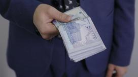 Мужчина в костюме держит в руке пачку денег номиналом 10 тысяч тенге