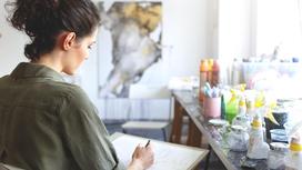 Девушка рисует простым карандашом на бумаге сидя у стола с красками