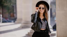 Девушка в темных брюках, кожаной куртке, полосатом топе, шляпе и солнцезащитных очках
