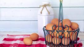 На столе лежит клетчатое кухонное полотенце. На полотенце стоит корзина с яйцами, лежат отдельно два яйца и стоит белая пластиковая бутылка, перевязанная веревкой