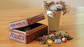 На столе стоит открытая деревянная шкатулка с резьбой, лажат рассыпанные камни-самоцветы и ваза с искусственными цветами