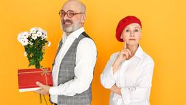Мужчина в очках и с бородой стоит к женщине спиной и держит в руках белые цветы и подарочную коробку. Женщина в красном берете стоит рядом, подперев щеку правой рукой