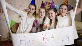 Четыре девочки в праздничных колпачках держат в руках плакат с поздравительной надписью на английском языке. Они радостно поздравляют одноклассницу с днем рождения