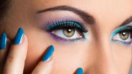Девушка с ярким макияжем глаз в голубых и лиловых оттенках трогает лицо рукой с синим маникюром