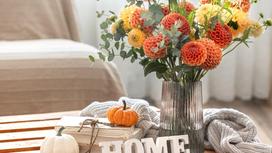 Осенние цветы в вазе стоят на журнальном столике. Рядом лежат две тыквы, книги, вязаный свитер, высечка  «Home»