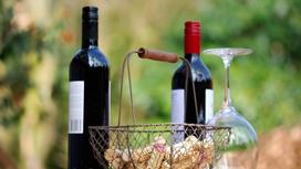 На столе стоят две темные бутылки с вином, металлическая корзинка с корковыми пробками, перевернутый винный бокал
