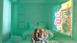 Две девочки подростки сидят на полу в комнате и смотрят на большой экран на стене, где идет фильм