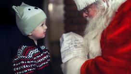 Мальчик в шапке с ушками и свитере разговаривает с Дедом Морозом. Дед Мороз в очках и в красной шубе держит в руках подарок для мальчика