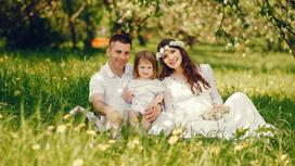 На зеленой лужайке сидит папа, мам и маленькая дочка. Они одеты в белые наряды, а у мамы на голове венок из белых цветов