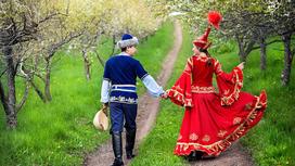 Парень с девушкой в национальных казахских костюмах идут по дороге в саду, держась за руки. Парень держит в руке домбру