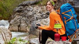 Девушка с туристическим рюкзаком и фотоаппаратом присела на камне у реки