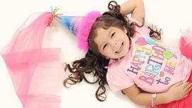Девочка с длинными курчавыми волосами в праздничном колпачке с розовым шлейфом, розовой футболке с поздравительными принтами лежит и улыбается.
