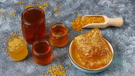 Мед разных цветов в открытых банках, соты с медом в мискуе, пыльца в деревянном совке и рассыпана на столе