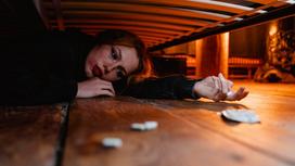 Девушка в темном свитере лежит на полу под кроватью с таблетками в руках