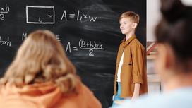 Школьник в коричневой рубашке стоит у доски, исписанной математическими формулами