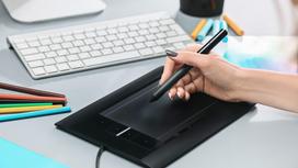 На столе лежит планшет, клавиатура, карандаши и маркеры. Девушка держит в руке стилус над планшетом