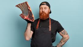 Мужчина в фартуке с рыжей бородой держит в правой руке на весу большой кусок мяса с ребром