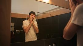 Унылый мужчина стоит у зеркала и смотрит на свое отражение, закрывая лицо руками