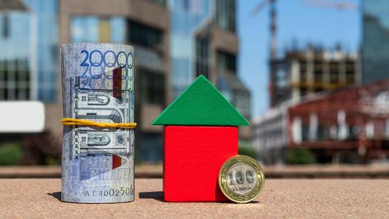 Свернутая купюра 20 тысяч тенге и монета 100 тенге стоят рядом с кубиками в виде дома Свернутая купюра 20 тысяч тенге и монета 100 тенге стоят рядом с кубиками в виде дома